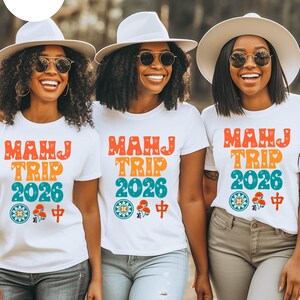 May include: Three women wearing white t-shirts with a colorful "MAHJ TRIP 2026" graphic, sunglasses, and white hats. The shirts have a floral and Chinese character design. They are smiling and wearing jeans.