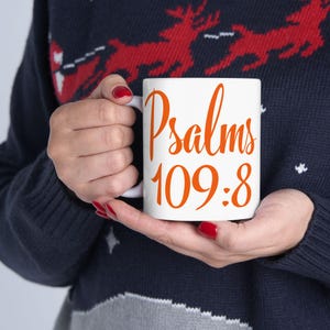 Puede incluir: Taza de cerámica blanca con el texto "Psalms 109:8" en escritura naranja. La taza la sostiene una persona que lleva un suéter azul marino con un diseño de reno rojo. La persona tiene esmalte de uñas rojo.