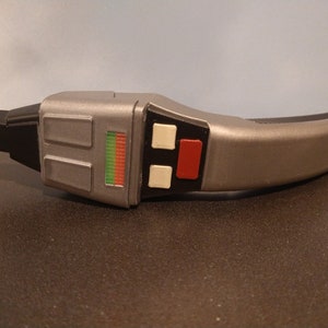 Peut inclure: Un appareil portatif argenté et noir avec un panneau de boutons rouge et blanc et une bande lumineuse verte et rouge. L'appareil est une réplique d'un phaser de Star Trek.