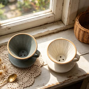 Könnte beinhalten: Zwei Keramik-Kaffeefilter mit Griffen, einer in gesprenkeltem Blaugrau und einer in gesprenkeltem Off-White. Beide verfügen über interne vertikale Rillen, einen runden Boden und drei Abflusslöcher sowie einen hellbraunen Randakzent.