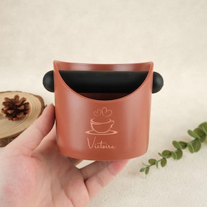 May include: A brown coffee grounds knock box with a black rubber bar and black side handles. The knock box features a coffee cup and heart design with the word "Victoire" printed on the front.