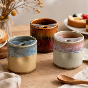 May include: Three cylindrical ceramic holders with a unique top shelf feature. One holder has a blue and tan glaze, one is a deep brown and amber, and the third features a pink and cream finish. Each piece has a small circular opening on the top ledge.