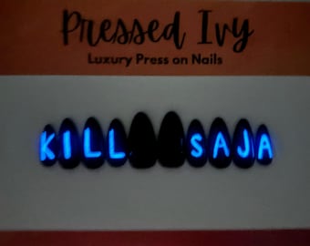 May include: Black press-on nails with the words "KILL SAJA" in glowing blue letters. The nails are almond-shaped and arranged on a white surface. The brand name "Pressed Ivy" is at the top.