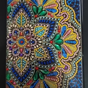 May include: A rectangular, decorative item with a detailed, colorful design. The design features a symmetrical pattern of flowers, leaves, and dots in shades of blue, green, yellow, red, and purple. The item has a black border and a glossy finish.
