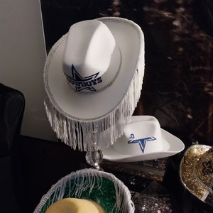 Może przedstawiać: Trzy białe kowbojskie kapelusze z frędzlami. G&oacute;rny kapelusz ma niebieską gwiazdę i napis "COWBOYS". Środkowy kapelusz ma niebieską gwiazdę. Dolny kapelusz ma zielono-ż&oacute;łty brzeg.