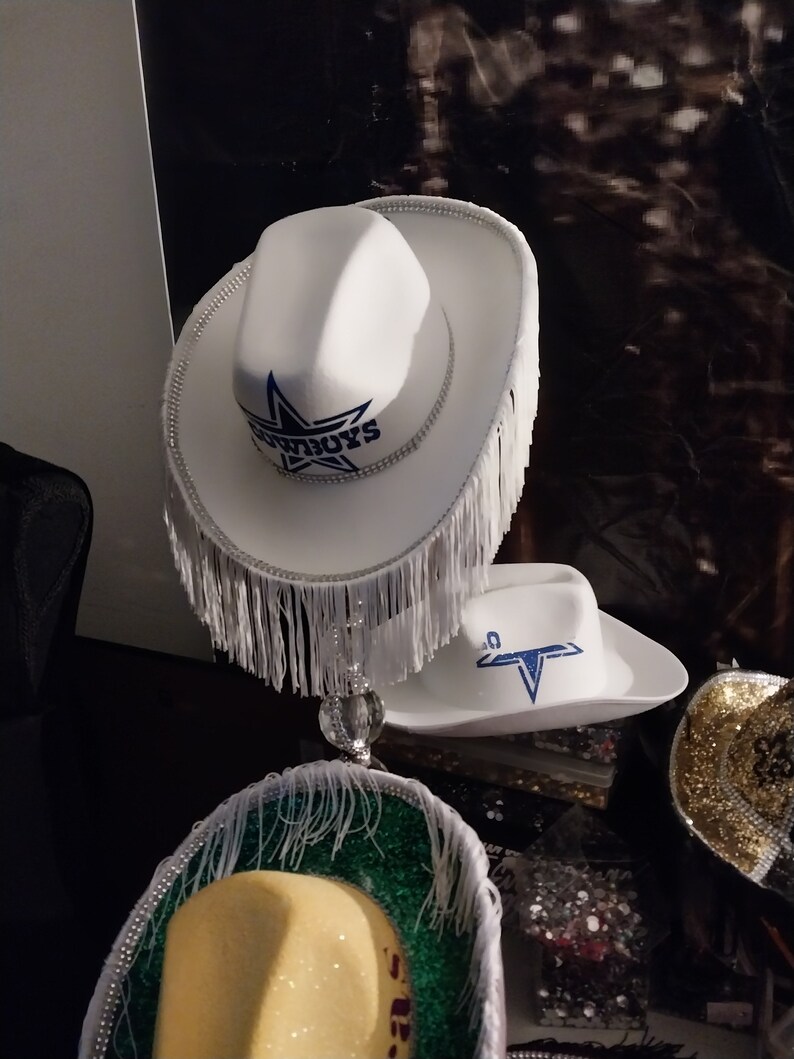 Może przedstawiać: Trzy białe kowbojskie kapelusze z frędzlami. G&oacute;rny kapelusz ma niebieską gwiazdę i napis "COWBOYS". Środkowy kapelusz ma niebieską gwiazdę. Dolny kapelusz ma zielono-ż&oacute;łty brzeg.