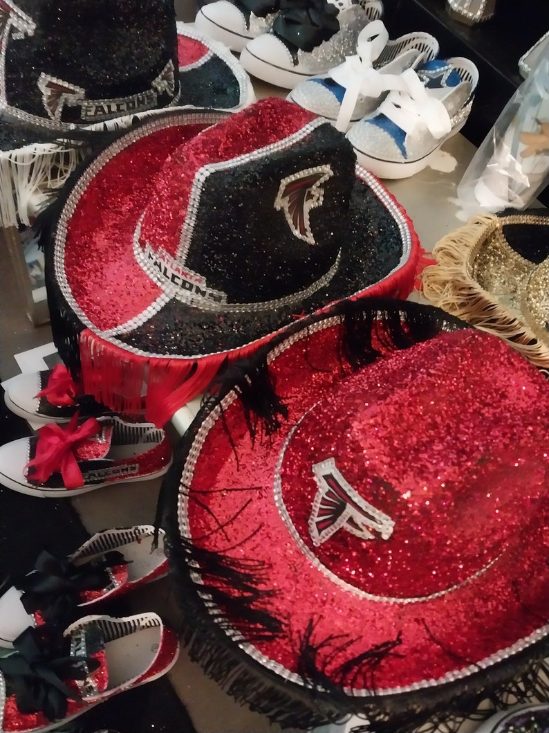 Może przedstawiać: Dwa czerwono-czarne kowbojskie kapelusze z czarnymi frędzlami i srebrnymi ozdobami. Kapelusze mają czerwoną i czarną powierzchnię z brokatem i białe logo Atlanta Falcons.