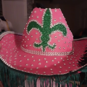 May include: Pink cowboy hat with a green glitter fleur-de-lis design and silver rhinestones.