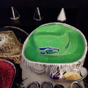 Może przedstawiać: Zielony i biały kapelusz kowbojski z logo Seattle Seahawks i kryształkami. Kapelusz ma biały frędzle wok&oacute;ł ronda.