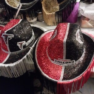 Może przedstawiać: Dwa czarne i czerwone kowbojskie kapelusze z srebrnymi frędzlami i kryształkami. Kapelusze mają napis "Atlanta Falcons".