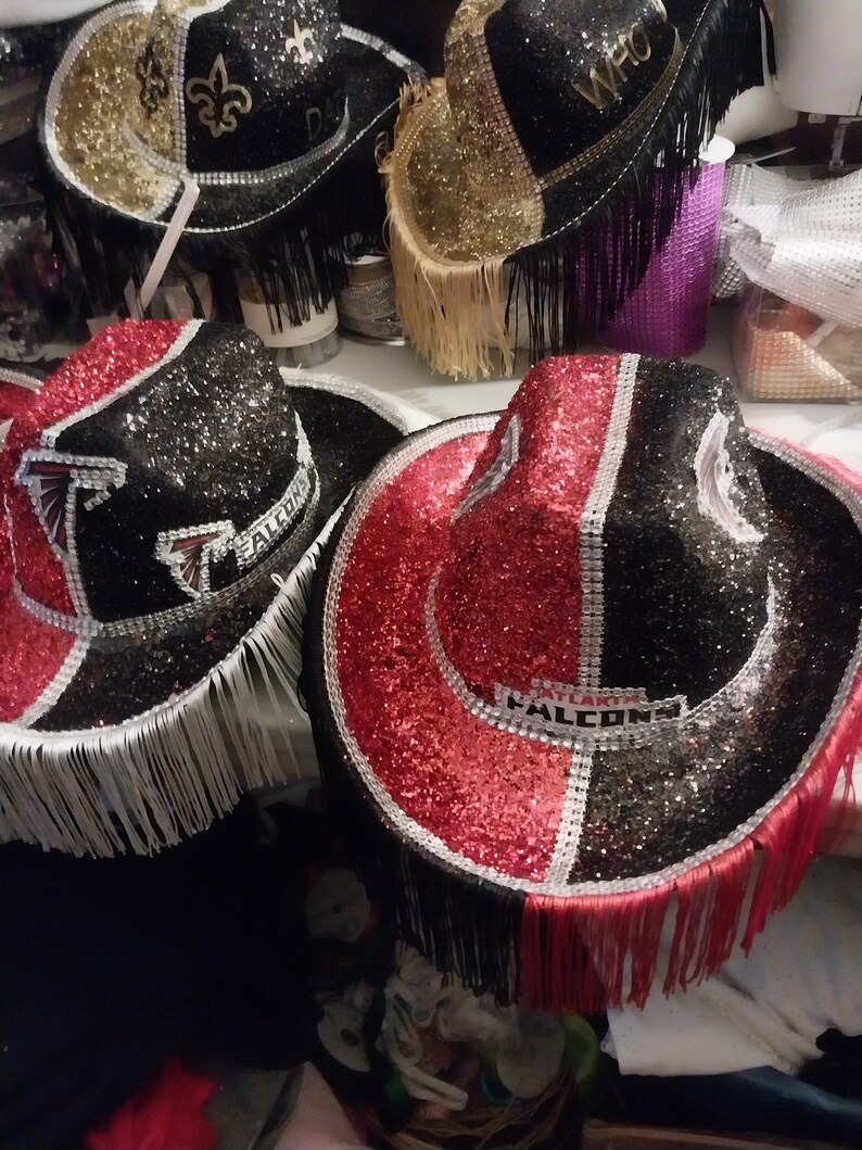 Może przedstawiać: Dwa czarne i czerwone kowbojskie kapelusze z srebrnymi frędzlami i kryształkami. Kapelusze mają napis "Atlanta Falcons".