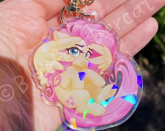 May include: A clear acrylic keychain featuring a cartoon pony with a pale yellow body, pink mane and tail, and feathered wings. The charm has a holographic finish with scattered iridescent shards and a gold-colored metal flower-shaped clasp.