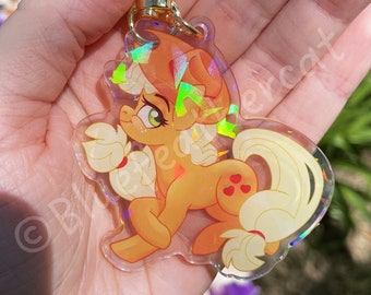 May include: An acrylic keychain featuring a cartoon pony with orange fur, a light yellow mane and tail, and a three-apple symbol on its flank. The charm has a holographic finish with geometric light reflections and a gold-colored star-shaped clasp.