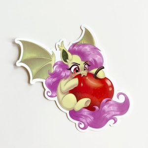 May include: A die-cut sticker featuring a pale yellow creature with purple hair and bat-like wings, biting into a large, shiny red apple. The creature has pointed ears, red eyes, and small fangs, depicted in a cartoon style with a white border.