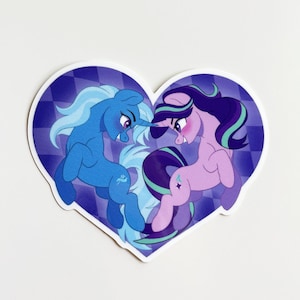 May include: A heart-shaped sticker featuring two cartoon ponies facing each other. The left pony is blue with a light blue mane and tail. The right pony is light purple with a dark purple and teal mane and tail. The background is a purple checkerboard.