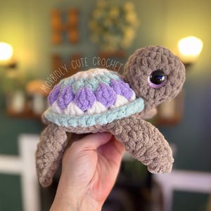 May include: A crocheted turtle toy with a purple, white and blue shell. The turtle has large, googly eyes and a brown body.