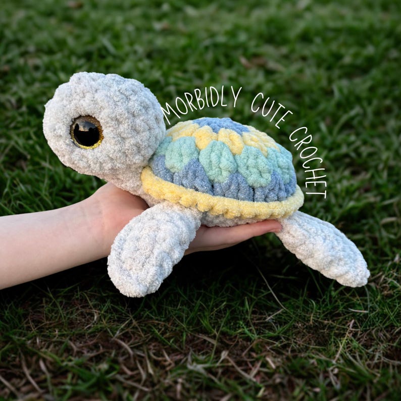 May include: A crocheted stuffed sea turtle toy with a light grey body, yellow and blue shell, and large brown eyes. The text "Morbidly Cute Crochet" is written in a white circle around the turtle.