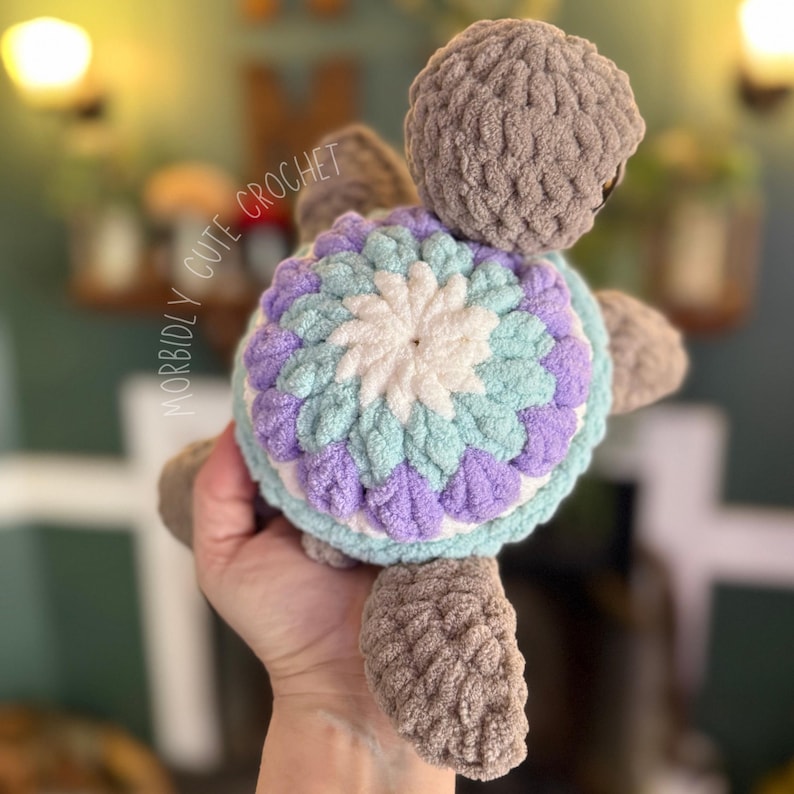 May include: A crocheted turtle toy with a purple, white and light blue shell. The turtle has a grey body and is held in a hand.