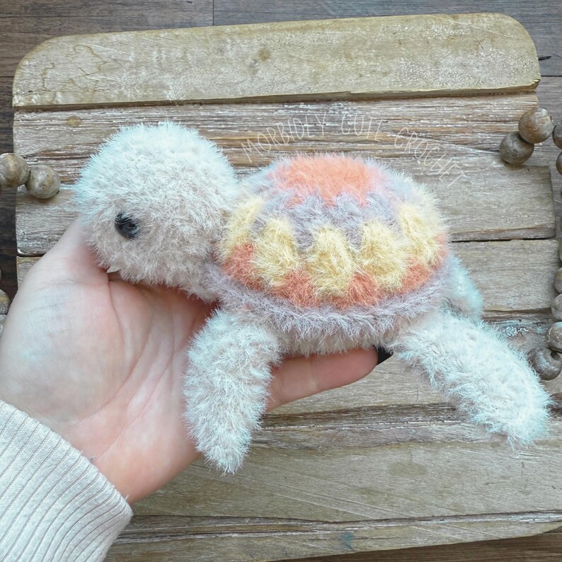 May include: A crocheted plush turtle toy with a pink, yellow and grey shell. The turtle is made with soft, fluffy yarn and has a sweet, friendly expression.