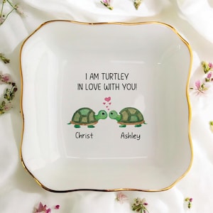 May include: White ceramic trinket dish with a gold rim. The dish features a watercolor design of two green turtles kissing, with the text "I AM TURTLEY IN LOVE WITH YOU!" and the names "Christ" and "Ashley".
