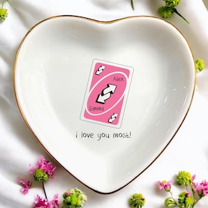 May include: Heart-shaped white ceramic dish with a gold rim. A pink Uno reverse card is in the center, with the names "Alex" and "Emma". The phrase "i love you most!" is below the card. Decorative flowers surround the dish.