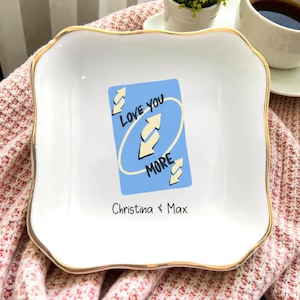 May include: A white ceramic trinket dish with a gold rim and a wavy edge. The dish features a blue card design with the words "LOVE YOU MORE" and the names "Christina & Max".