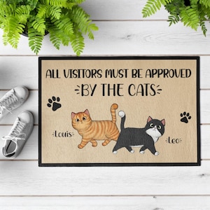 Puede incluir: Una alfombra rectangular con el texto "ALL VISITORS MUST BE APPROVED BY THE CATS". Presenta ilustraciones de dos gatos, un atigrado naranja llamado Louis y un gato blanco y negro llamado Leo, con huellas de patas.