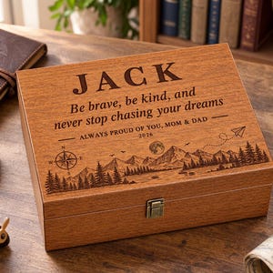 May include: A rectangular wooden keepsake box with a hinged lid and a metal latch. The lid is engraved with the name JACK, the text Be brave, be kind, and never stop chasing your dreams, ALWAYS PROUD OF YOU, MOM & DAD, 2026, and a mountain landscape.