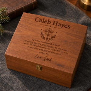 Op de afbeelding: Een rechthoekige houten aandenkendoos met een scharnierend deksel en een koperen sluiting. Het deksel is lasergegraveerd met de naam Caleb Hayes, een kruisicoon, het bijbelvers Jozua 1:9 en de handtekening Love, Dad.