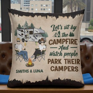 Puede incluir: Un cojín decorativo con temática de camping con borde negro. El cojín presenta una pareja sentada junto a una fogata, una autocaravana y el texto "Sentémonos junto a la fogata y veamos a la gente estacionar sus autocaravanas". Los nombres "Smiths & Luna" también están impresos.