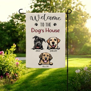 Puede incluir: Una bandera de jardín beige con el texto "Welcome to the Dog's House" y huellas de patas. La bandera presenta ilustraciones de tres perros con los nombres Jazz, Shelby y Ollie.