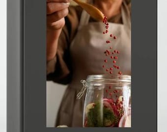May include: A book cover with a photo of a person adding red peppercorns to a jar of pickled vegetables. The text on the cover reads "Homestead Kitchen: 12 Homemade Staples to Master in a Year".