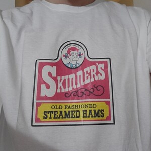 May include: White t-shirt with a graphic of a vintage food label. The label reads "Skinner's Old Fashioned Steamed Hams" in red and yellow, with a cartoon character illustration at the top. The shirt is made of a soft, comfortable material.