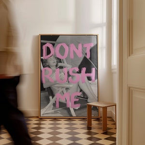 Könnte beinhalten: Gerahmter Schwarz-Weiß-Druck mit einer Frau in einem Liegestuhl, überlagert mit rosa Text "DON'T RUSH ME". Das Kunstwerk befindet sich in einem Raum mit Schachbrettmuster und einem kleinen Holzschemel, was eine entspannte Atmosphäre schafft.