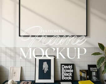 May include: A black framed picture mockup with a white background. The frame is hanging on a white tiled wall. There are several other framed pictures and books on a blue shelf below the frame.