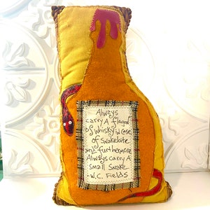 May include: A yellow felt pillow shaped like a bottle with a red felt top. The pillow has a small red felt snake on the side and a white label with the text "Always carry a flagon of whisky in case of Snakebite and furthermore Always carry a small Snake. -W.C. Fields"