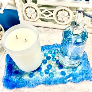 May include: A blue and white resin tray with a textured surface and a pattern of blue and white glass beads. A white frosted glass candle and a clear glass bottle with a silver pump dispenser are sitting on the tray.