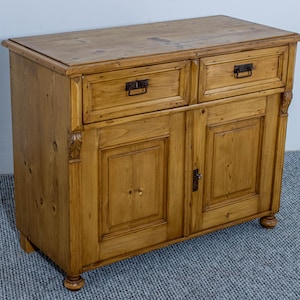 Può includere: Un mobile in legno marrone chiaro con due cassetti e due ante. Il mobile ha una forma rettangolare con un piano leggermente sporgente. I cassetti e le ante hanno maniglie in metallo scuro. Il mobile è in legno con venature visibili.