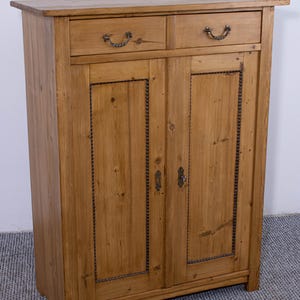 May include: A light brown wooden cabinet with two drawers and two doors. The cabinet has decorative metal handles and trim. The wood grain is visible throughout the cabinet.