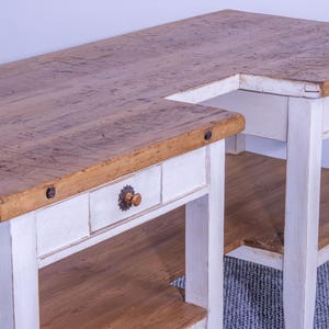 Peut inclure: Table en bois rustique en forme de L, avec une finition vieillie. Le plateau est brun clair avec le grain du bois visible, et la base est peinte en blanc. Comprend des tiroirs avec des poignées ornées et une étagère inférieure.