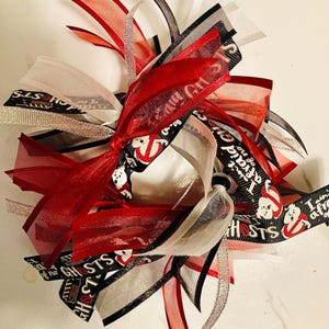 Op de afbeelding: Een decoratieve strik met rode, zwarte, witte en zilveren linten. Sommige linten hebben tekst en spookafbeeldingen. De strik is in het midden geknoopt en heeft meerdere streamers.