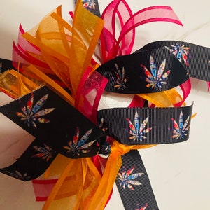 Könnte beinhalten: Eine bunte Schleife mit einem schwarzen Band, das ein sich wiederholendes Muster aus bunten Marihuana-Blättern zeigt. Die Schleife enthält auch ein durchsichtiges rotes und orangefarbenes Band.