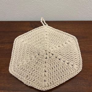 May include: A beige, hexagonal crochet potholder with a loop for hanging. The handmade kitchen item features a textured, floral-like design in the center and a solid border. The potholder is made of a soft, natural-looking yarn.