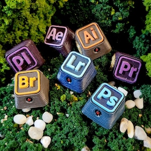 May include: A collection of colorful, square keycaps featuring the logos for Adobe software, including Ae, Ai, Pr, Ps, Lr, Pl, and Br. Each keycap has a unique color scheme, with a small red accent. The keycaps are set against a backdrop of green foliage.