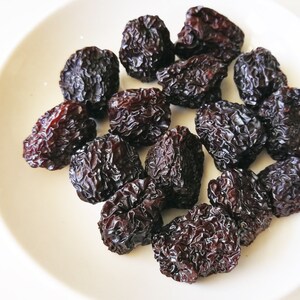 May include: A close-up shot of a collection of dark, wrinkled dried plums. The plums are a deep reddish-brown color and have a textured surface. They are arranged on a white plate, creating a simple and natural presentation.
