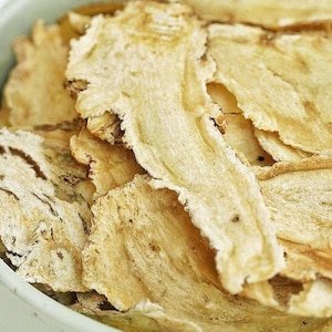 May include: Close-up of dried, light beige slices of a root vegetable, possibly used in herbal medicine. The slices have a slightly rough texture and varying shapes, with some showing darker brown edges. The image is well-lit, highlighting the natural colors and textures.