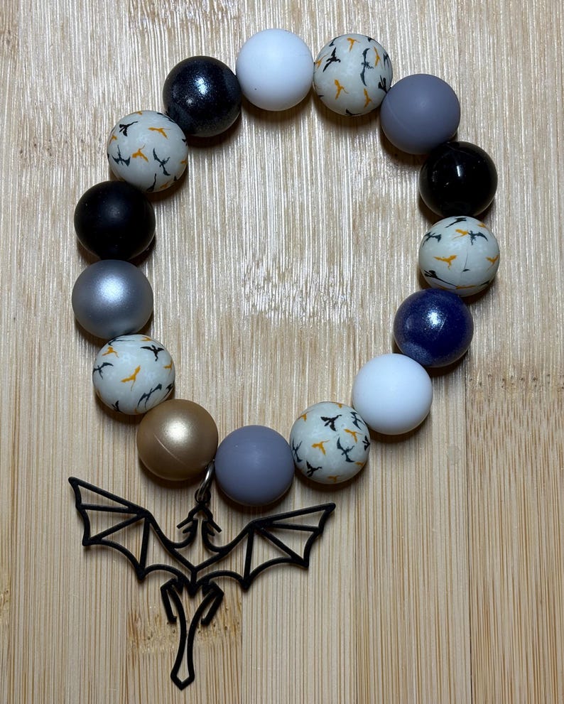 Op de afbeelding: Een armband gemaakt van kleurrijke kralen met een zwarte drakenbedel. De kralen zijn een mix van zwart, wit, grijs, zilver, goud en blauw. Sommige kralen hebben een patroon van kleine zwarte vogels op een witte achtergrond.
