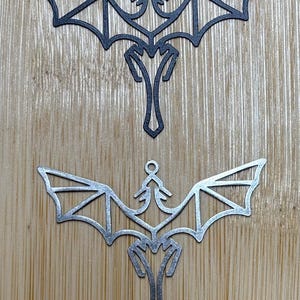 Op de afbeelding: Twee metalen draken ornamenten, een zwart en een zilver, met een geometrisch ontwerp. De ornamenten hangen aan een houten oppervlak.