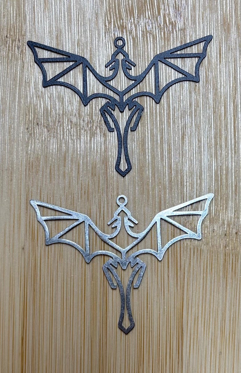 Op de afbeelding: Twee metalen draken ornamenten, een zwart en een zilver, met een geometrisch ontwerp. De ornamenten hangen aan een houten oppervlak.