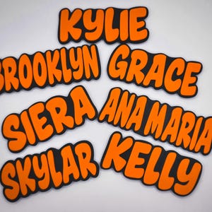 May include: Orange and black name cutouts. The names "Kylie", "Brooklyn", "Grace", "Siera", "Ana Maria", "Skylar", and "Kelly" are displayed in a bubble font with a black outline.
